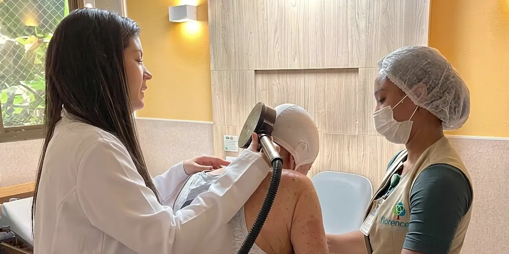 A Dra. Luana Amorim aplica a bobina de estimulação na cabeça de um paciente, de costas, enquanto a fisioterapeuta o apoia pelo braço