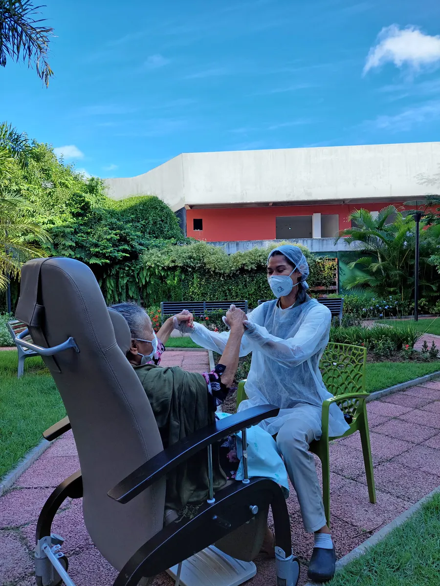 Profissional da equipe conduz mobilização de braços com paciente idosa em poltrona, no jardim da unidade.